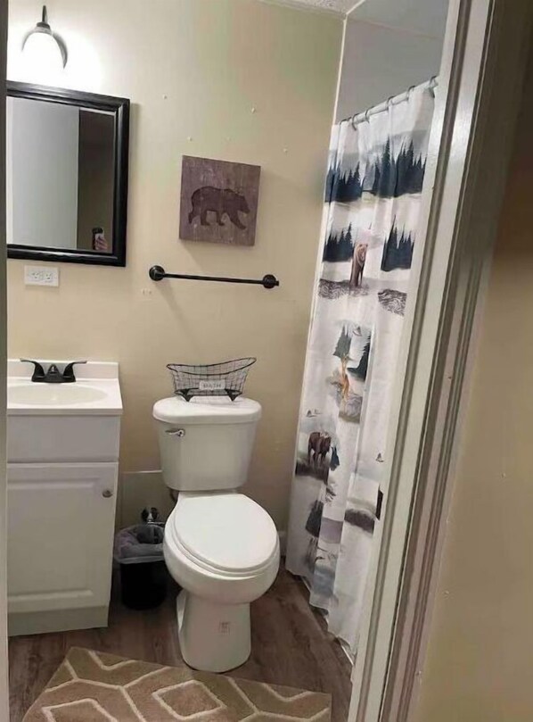 Une petite salle de bain avec des toilettes, un lavabo et un miroir. Un ours décoratif est accroché au mur, et le rideau de douche est orné d’un motif de forêt.