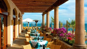 An outdoor dining area with tables and chairs, a view of the ocean, and a covered walkway with columns. - Room with a view of the Pacific and a private Jacuzzi at Sunset Beach Resort (Cabo San Lucas)