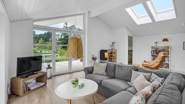 Ein modernes Wohnzimmer mit einem großen grauen Sofa, einem weißen Couchtisch und einem Fernseher auf einem Holzständer. Ein Bücherregal mit Büchern und ein kleiner Tisch mit Dekorationselementen sind ebenfalls zu sehen. Außerdem sind ein Korbstuhl und ein gepolsterter Sessel erkennbar.