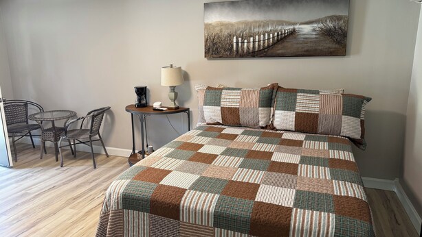 A bedroom with a large bed, a small table with a lamp, and a chair. There is a painting on the wall above the bed. - Motel at KY Lake. Reel & Relax (Gilbertsville)
