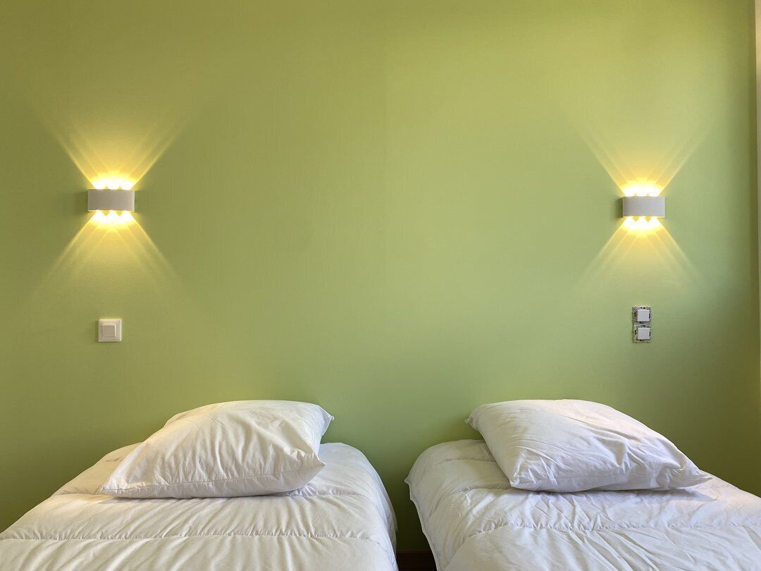 Ein Zimmer mit zwei Betten, jeweils mit weißer Bettwäsche und Kissen, an einer grünen Wand. An beiden Seiten der Betten befinden sich jeweils eine Wandleuchte.