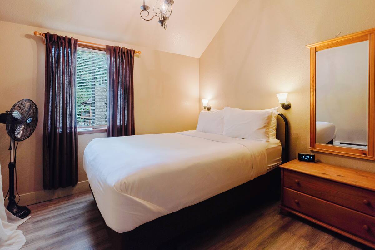 A hotel room with a bed, a nightstand, a mirror, a fan, and a window with curtains.