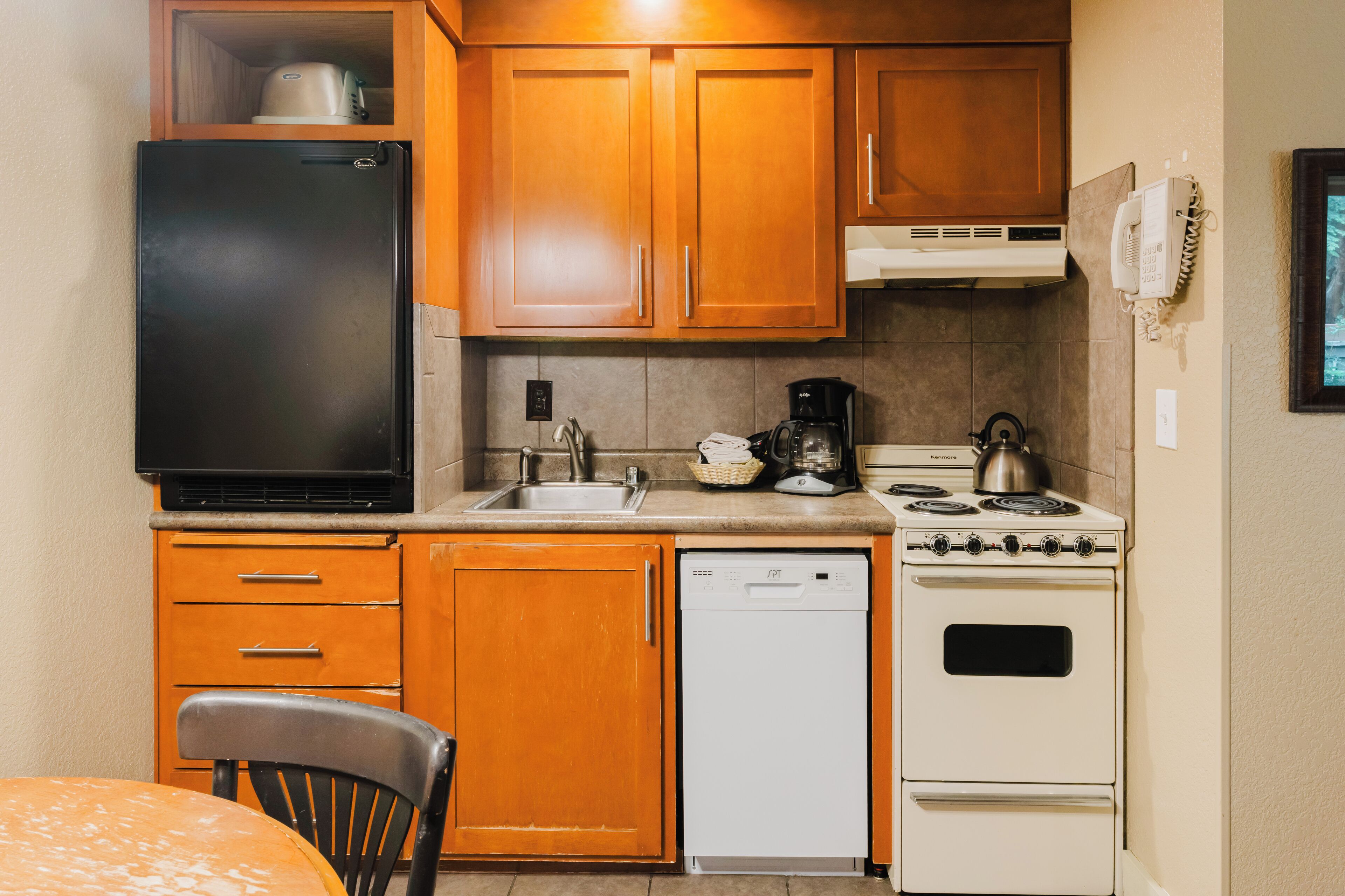 Une cuisine compacte avec des meubles en bois, un réfrigérateur noir, un lave-vaisselle blanc, une cuisinière et une machine à café.