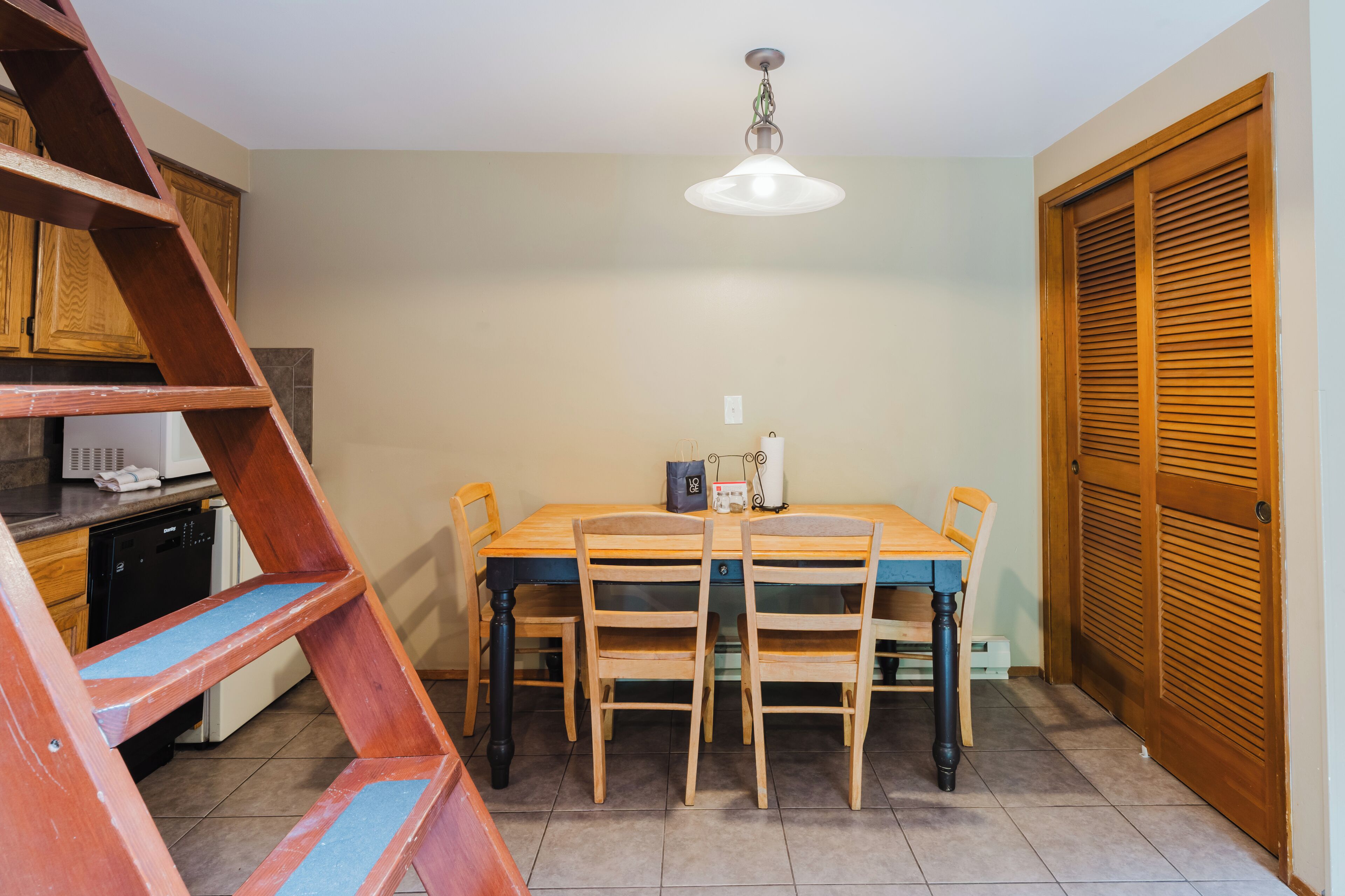 Un comedor con una mesa de madera, cuatro sillas, una lámpara en el techo, una escalera de madera y una puerta de madera.