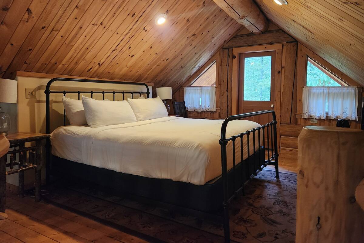 A wooden cabin bedroom with a bed, two bedside lamps, a window with curtains, and a wooden floor.