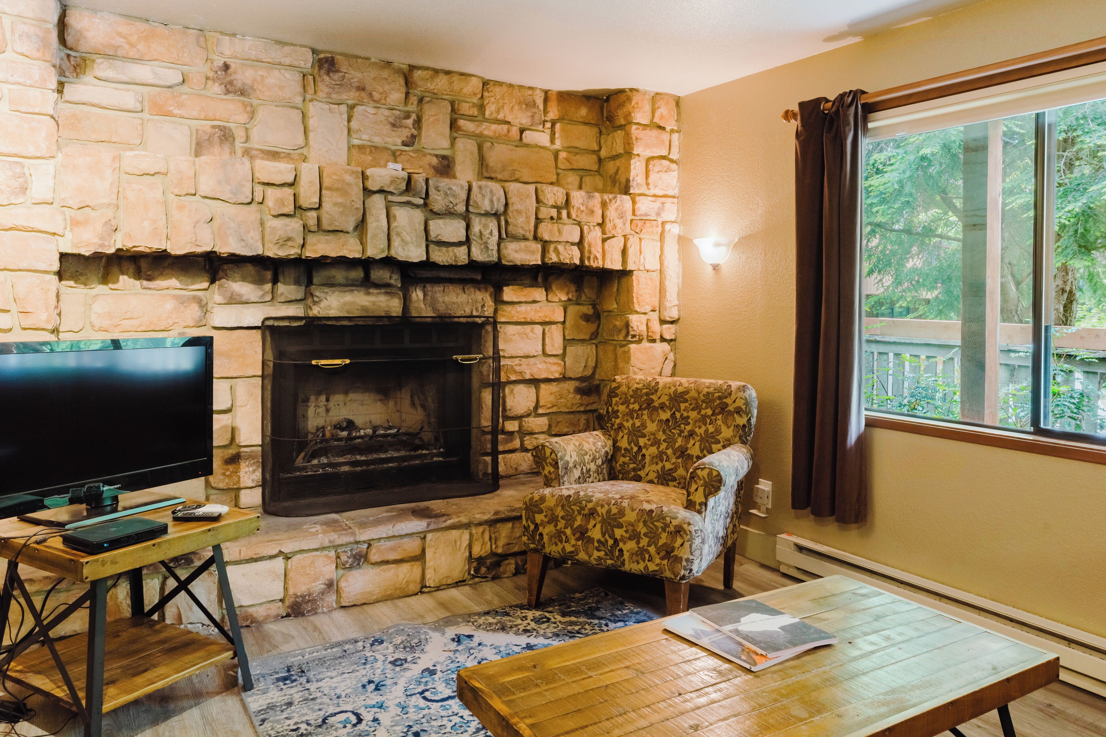 Una chimenea de piedra, un televisor, un sillón estampado y una mesa de centro de madera en una habitación con ventana y vista a la vegetación.