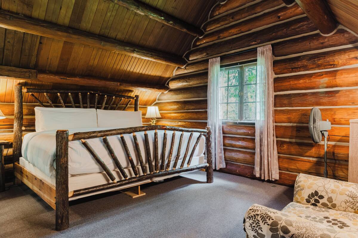 A log cabin bedroom with a wooden bed, a nightstand, a lamp, a window with curtains, and a fan.
