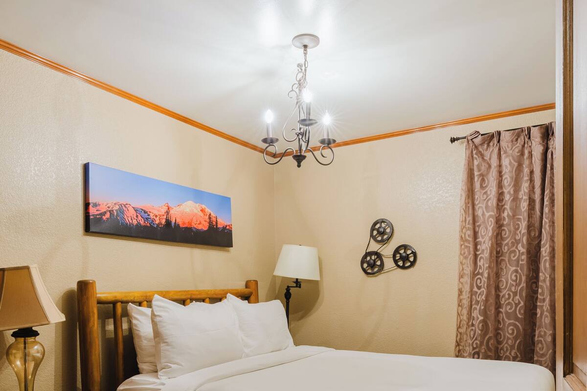 A neatly made bed with a wooden headboard, a nightstand with a lamp, a wall-mounted picture, and a chandelier.