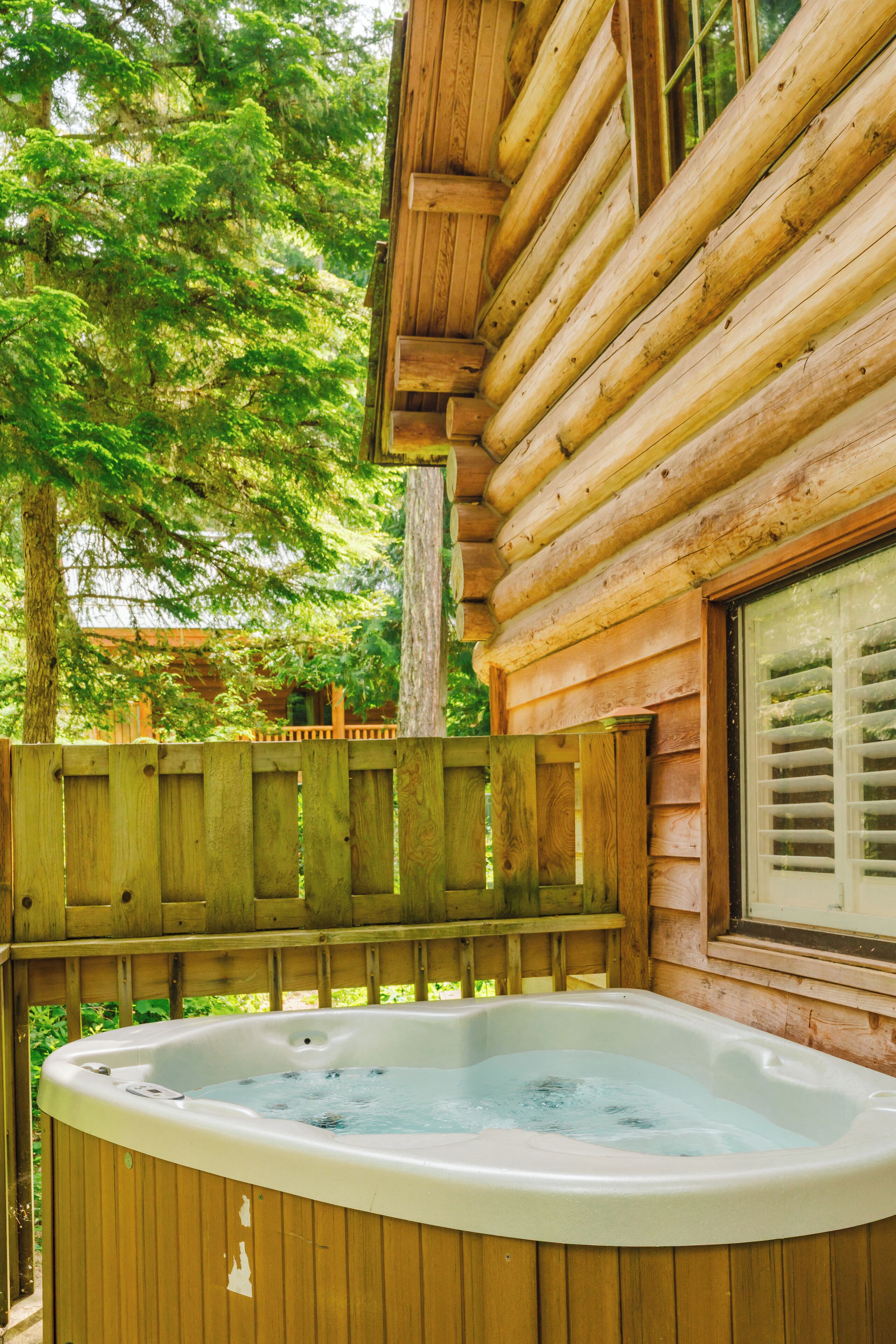 A wooden cabin with a hot tub in front of a wooden fence and a large tree.
