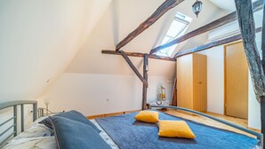 Ein Schlafzimmer im Loft-Stil mit freiliegenden Holzbalken, einem Bett mit blauer Bettwäsche und einem großen Fenster, das natürliches Licht hereinlässt.