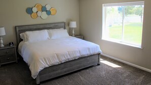 Une chambre à coucher comprenant un lit, des tables de chevet, une lampe, un élément décoratif mural et une fenêtre donnant sur une pelouse.