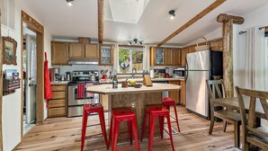 A kitchen with wooden cabinets, a central island with stools, a refrigerator, and a window above the sink. - 'RIVER HAVEN' is a serene riverside cabin on the Rogue River (Trail)