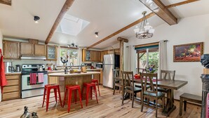 A spacious kitchen with wooden cabinets, a central island with stools, a dining area with a table and chairs, and a large window with a view of greenery. - 'RIVER HAVEN' is a serene riverside cabin on the Rogue River (Trail)