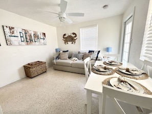 Uma sala de estar com um sofá, uma mesa de jantar posta com jogos americanos, um ventilador de teto, uma decoração de caranguejo na parede e uma placa com tema praiano.