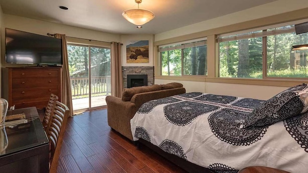 A bedroom with a large bed, a fireplace, a TV, and a balcony with a view of trees.
