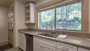 A kitchen with a granite countertop, white cabinets, a stainless steel sink, and a window with a view of trees.