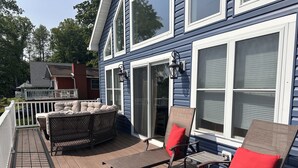 Eine Terrasse mit Gartenmöbeln, darunter eine Couch und zwei Stühle, mit Blick auf ein Haus mit großen Fenstern und einem weißen Geländer.