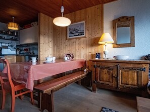 Una cocina acogedora con paredes de madera, una mesa de comedor con mantel a cuadros y un banco. Hay dos lámparas colgantes, una radio y un cuadro enmarcado en la pared.