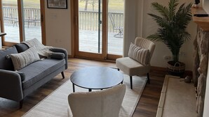 Un salón amplio con un gran ventanal, un sofá gris, un sillón blanco y una mesa de centro redonda.