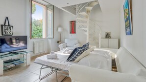 A modern hotel room with a bed, a TV, a glass coffee table, a white sofa, a spiral staircase, and a window with a view of greenery. - Appartement Perrissol - Welkeys (Cannes)
