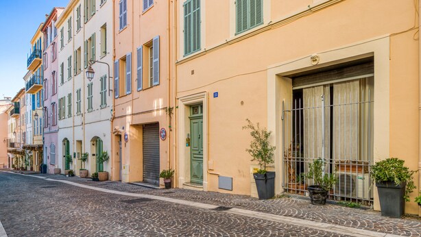 A cobblestone street lined with colorful buildings, each with shutters and potted plants. - Appartement Perrissol - Welkeys (Cannes)