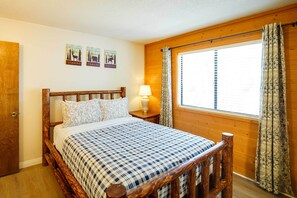Een bed met houten frame, een geruit beddegoed, een bedlamp en een raam met gordijnen.