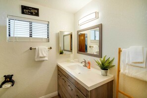 Een badkamer met een witte wasbak, houten wastafelmeubel, een spiegel, een handdoekrek en een bordje met gratis zeep en water.