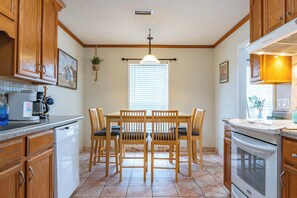 Une cuisine avec des meubles de rangement en bois, une table à manger avec des chaises, un four encastré et un luminaire suspendu.