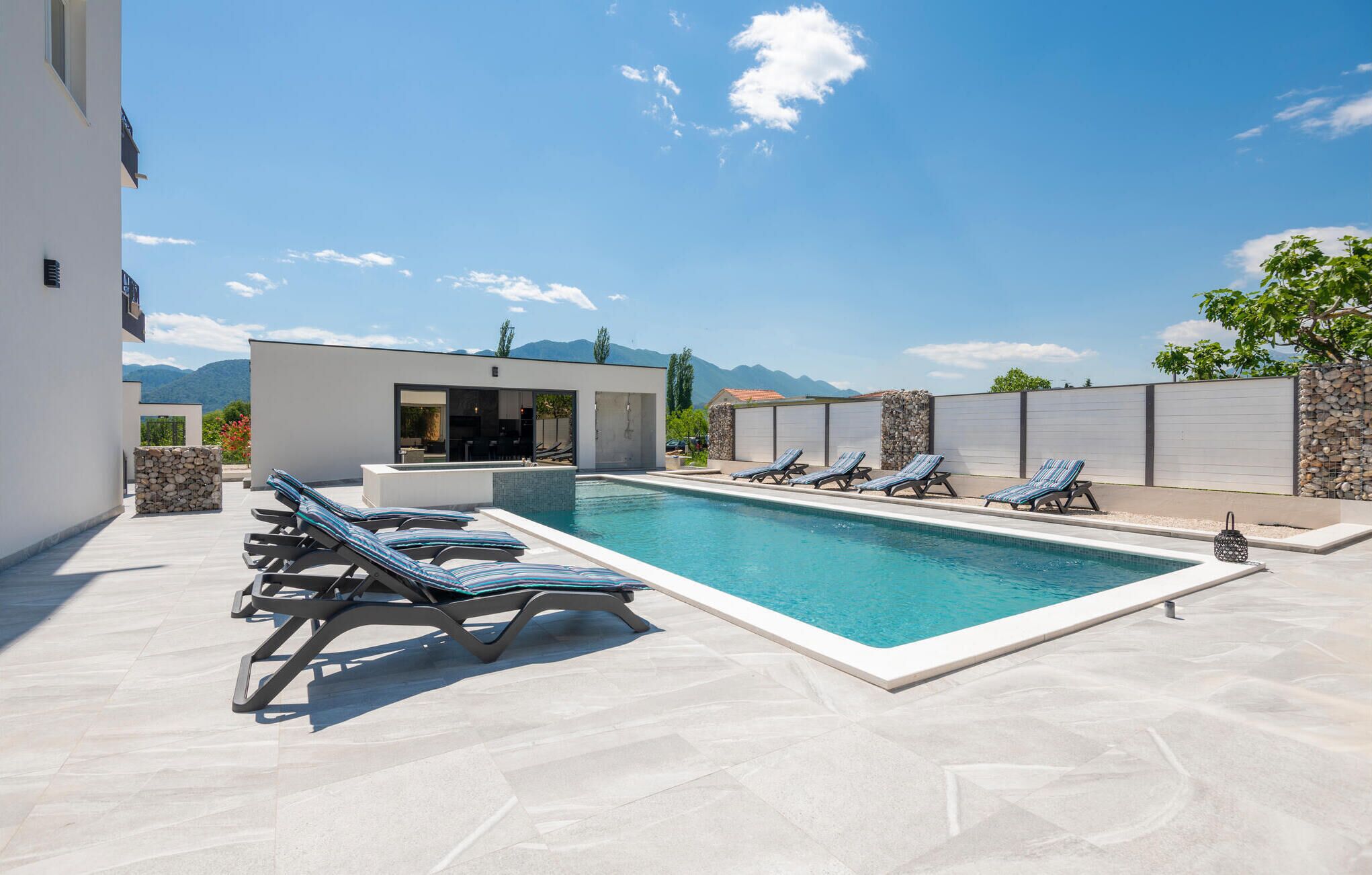 Un espace piscine extérieur moderne, avec des chaises longues, un petit bâtiment doté d’une porte en verre et une vue sur les montagnes en arrière-plan.