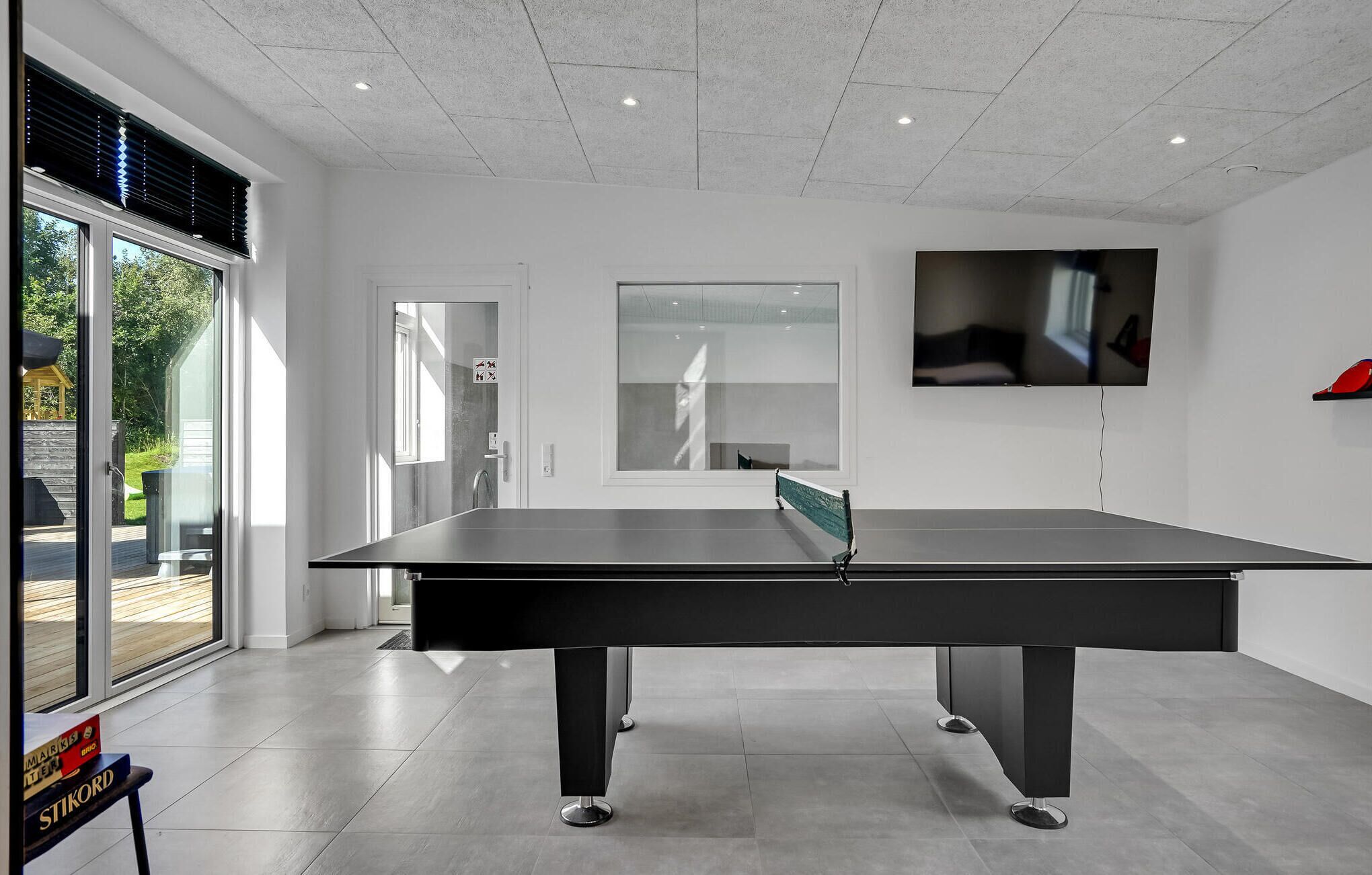 A modern indoor ping pong table with a glass barrier, a TV mounted on the wall, and a view of the outdoors through large windows.