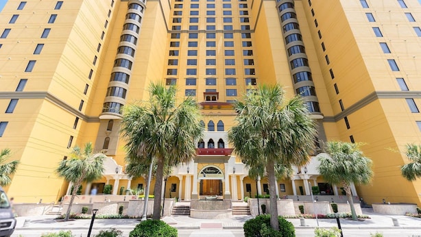 Un hotel de varios pisos con una entrada central, palmeras y cielo despejado.