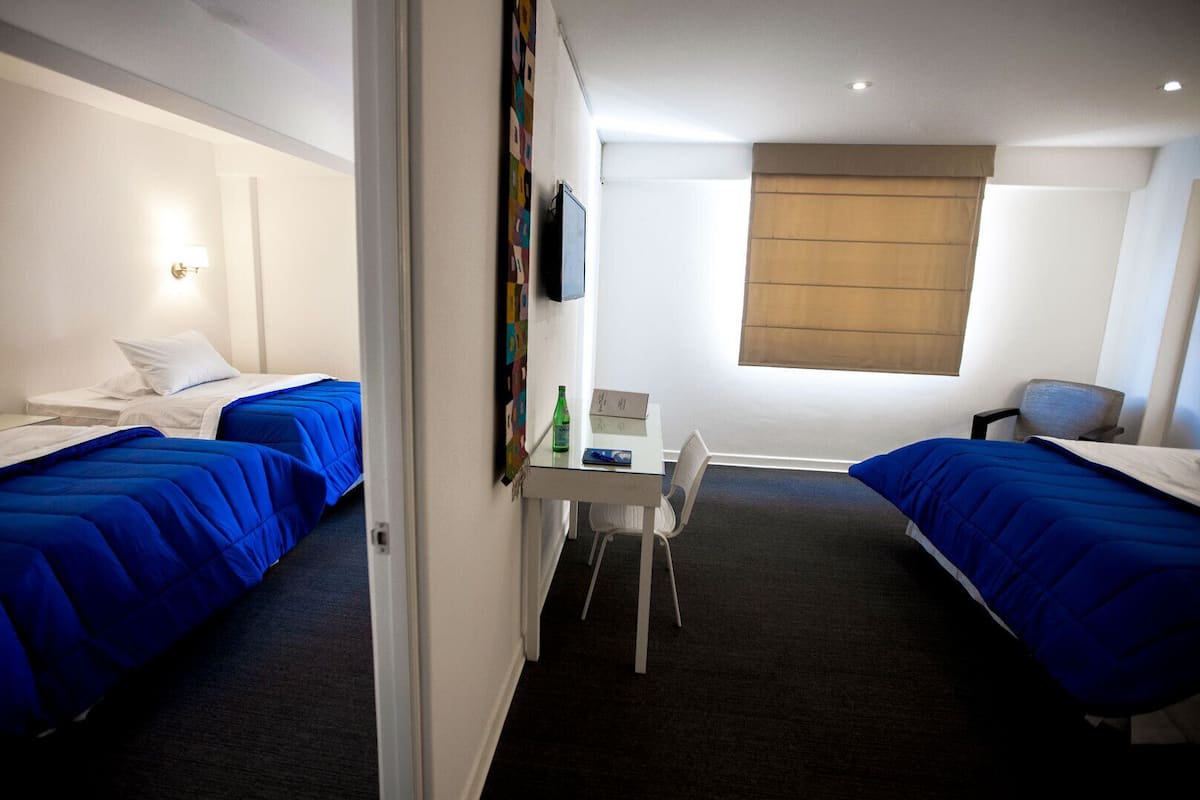 Ein Hotelzimmer mit zwei Betten, einem kleinen Schreibtisch, einem Stuhl und einem Fenster mit Jalousien.