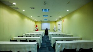 Un salón de conferencias con filas de mesas y sillas, una pantalla para proyector y un atril al frente.