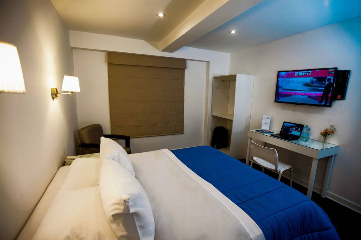 Ein Hotelzimmer mit einem Bett, einem Schreibtisch mit Laptop, einem Fernseher und einem Stuhl.