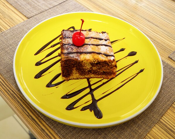 A slice of cake with chocolate drizzle and a cherry on a yellow plate. - Hotel Gran Palma Huancayo (El Tambo)
