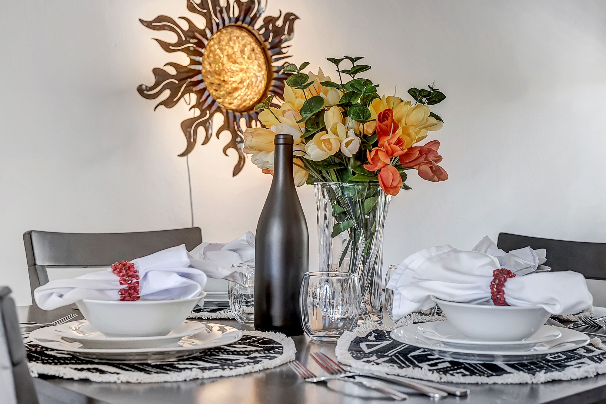 Ett matbord dukat med vita tallrikar, skålar och bestick, en genomskinlig glasvas med blommor och en dekorativ lampa formad som en sol.
