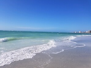 A beach with clear blue water, gentle waves, and a distant shoreline with buildings.