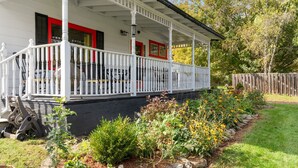 Un porche blanco con detalles rojos, una valla de madera y un jardín con diversas plantas y flores.