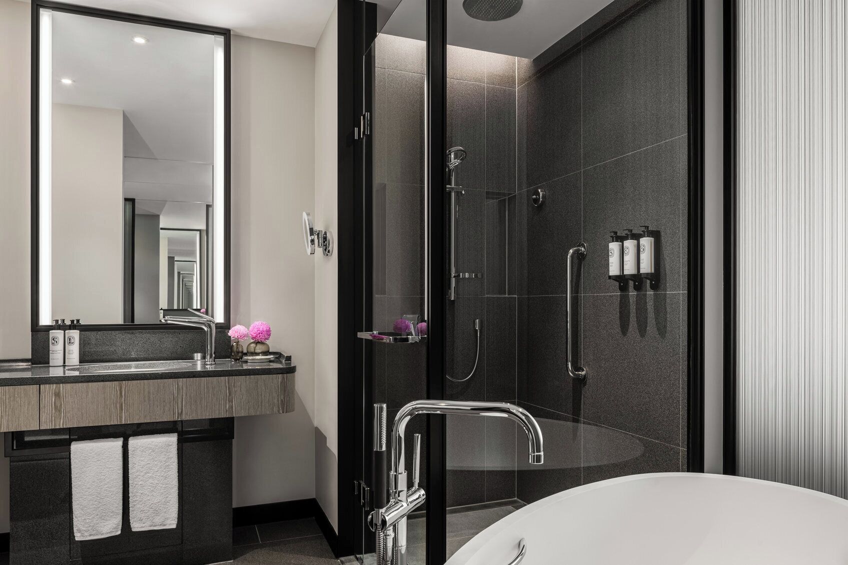 A modern bathroom with a large mirror, a freestanding bathtub, a glass-enclosed shower, and a vanity with a dark countertop and two towels.