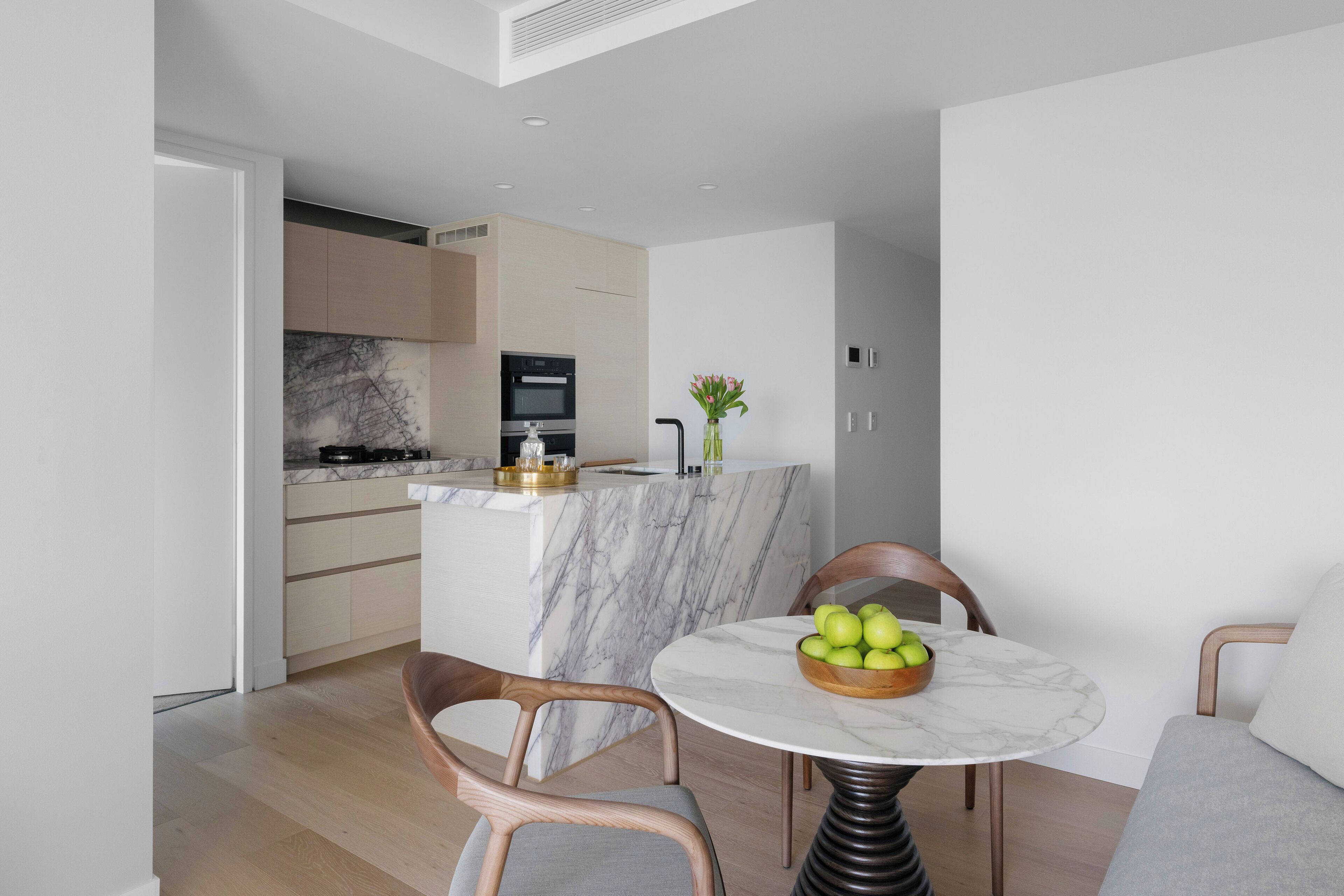 A modern kitchen with a marble island, wooden chairs, and a table with a bowl of apples.
