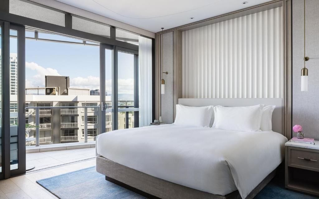 Une chambre d’hôtel moderne dotée d’un grand lit, d’une table de chevet et offrant une vue sur la ville grâce à de grandes fenêtres du sol au plafond.