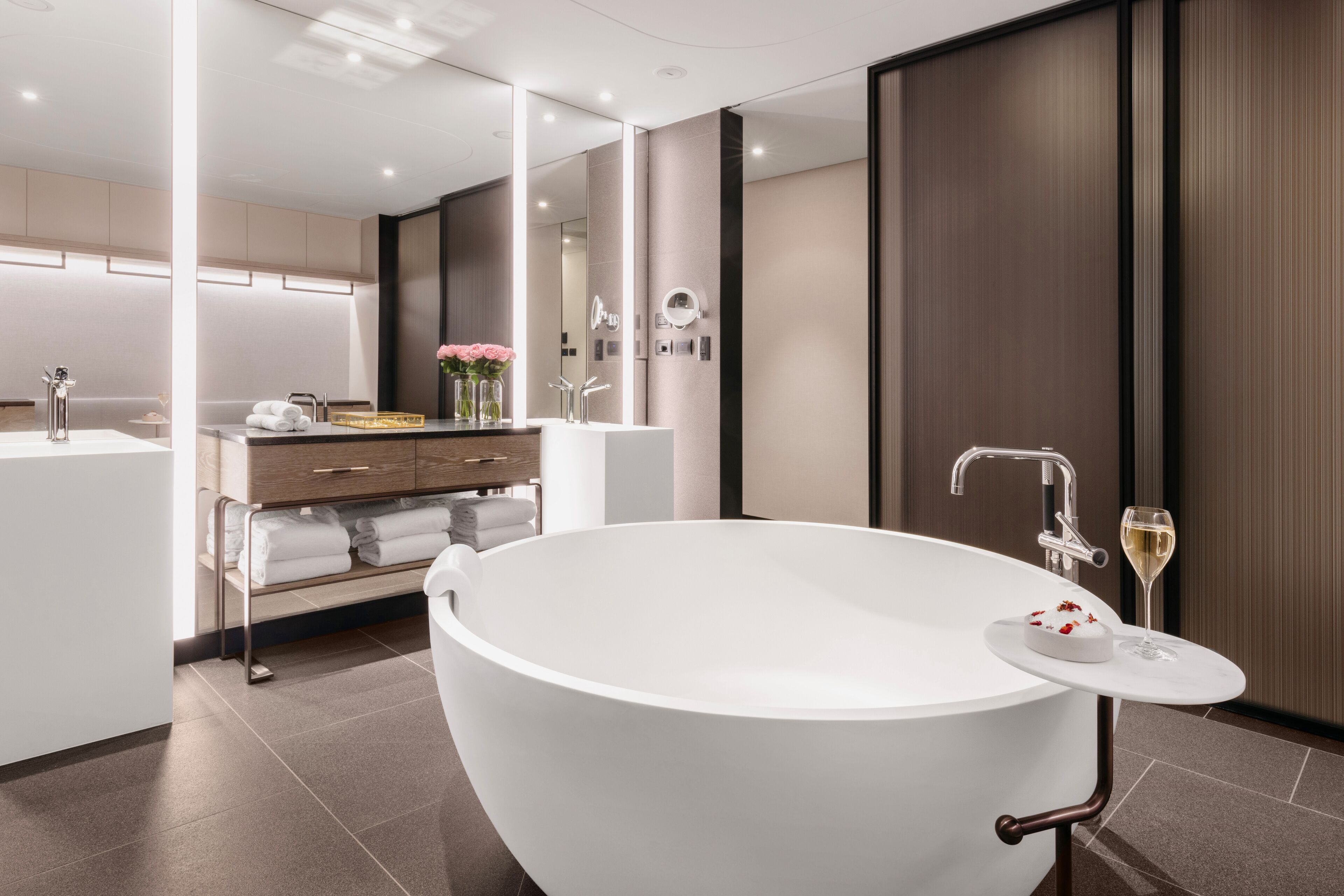 A modern bathroom with a freestanding bathtub, a vanity with a mirror, and a glass of wine on a small table.