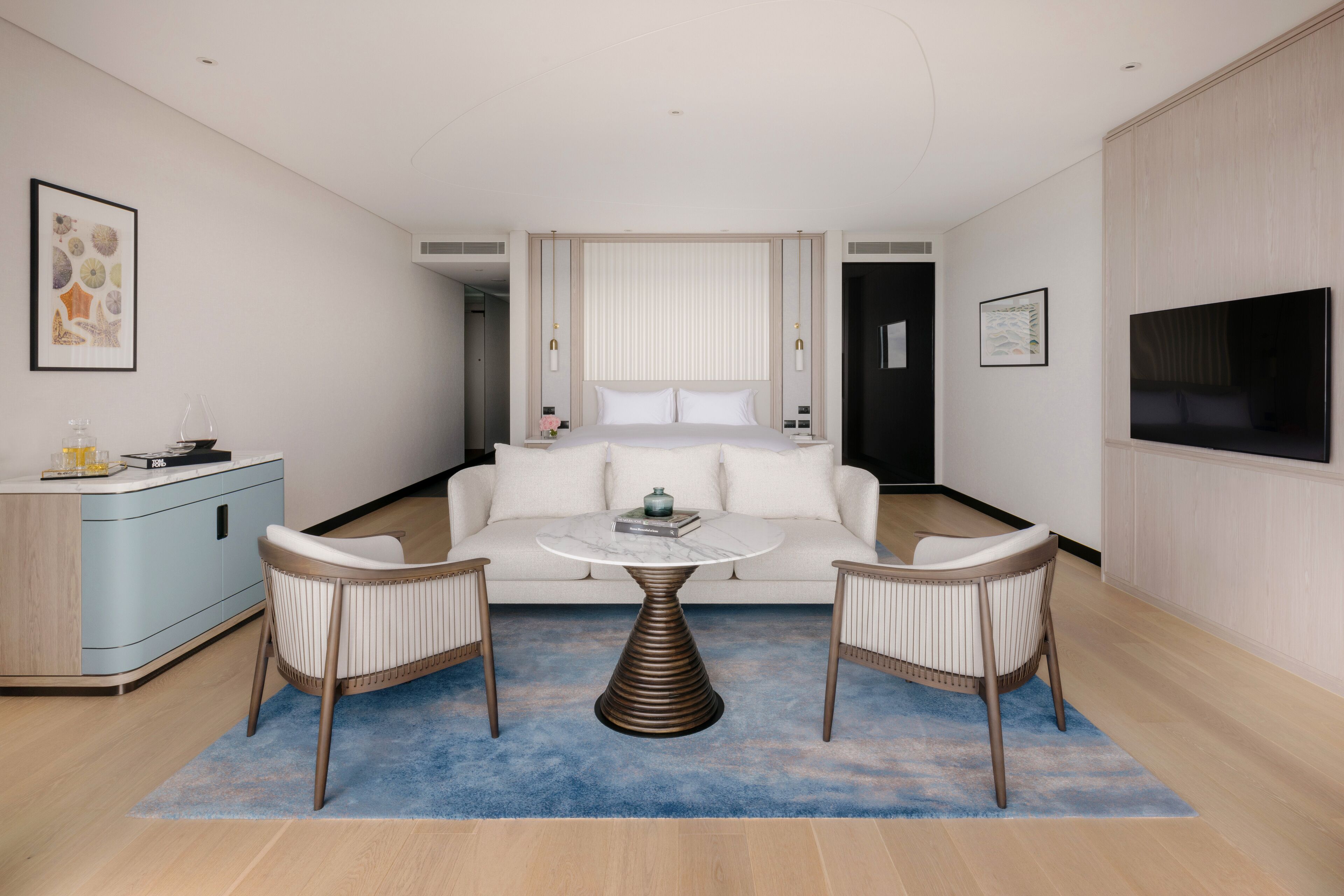 A modern hotel room with a central seating area, a sofa, two chairs, a coffee table, a TV, and artwork on the walls.
