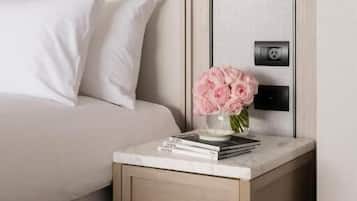 A neatly made bed with white linens, a bedside table with a drawer, a vase with pink flowers, and a wall-mounted light fixture.