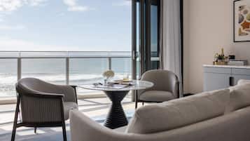 A modern room with a view of the ocean, featuring a round table, two chairs, a sofa, and a small side table with a bottle and glasses.