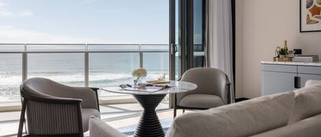 A modern room with a view of the ocean, featuring a round table, two chairs, a sofa, and a small side table with a bottle and glasses.
