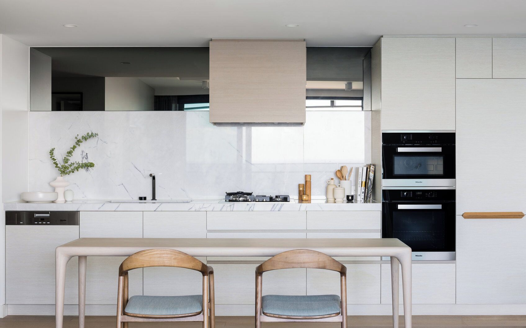 A modern kitchen with white cabinetry, a built-in oven, and a dining area with two wooden chairs.