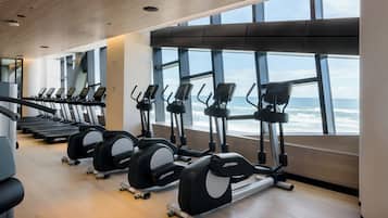 A gym with treadmills and elliptical machines, large windows with a view of the ocean, and wooden flooring.