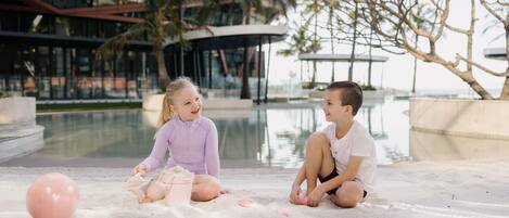 Dos niños jugando en una zona arenosa cerca de un edificio moderno con ventanas de vidrio y palmeras.