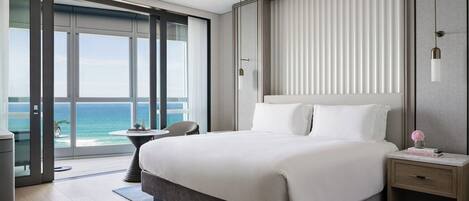 A modern hotel room with a large bed, a small table, a chair, and a view of the ocean through floor-to-ceiling windows.